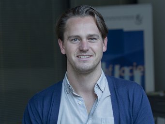 Ein Mann trägt ein weißes Hemd und einen blauen Cardigan. Im Hintergrund befindet sich ein unscharfes Plakat mit blauen und weißen Elementen.