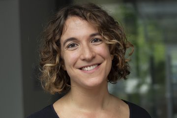 Individual with shoulder-length curly hair in a black shirt, situated indoors against a blurred backdrop of windows and greenery.