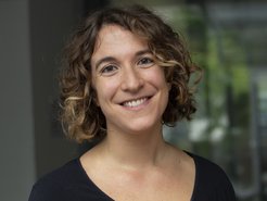 Individual with shoulder-length curly hair in a black shirt, situated indoors against a blurred backdrop of windows and greenery.