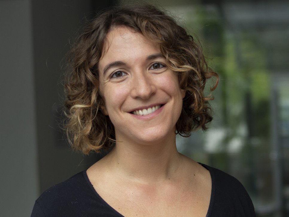 Individual with shoulder-length curly hair in a black shirt, situated indoors against a blurred backdrop of windows and greenery.