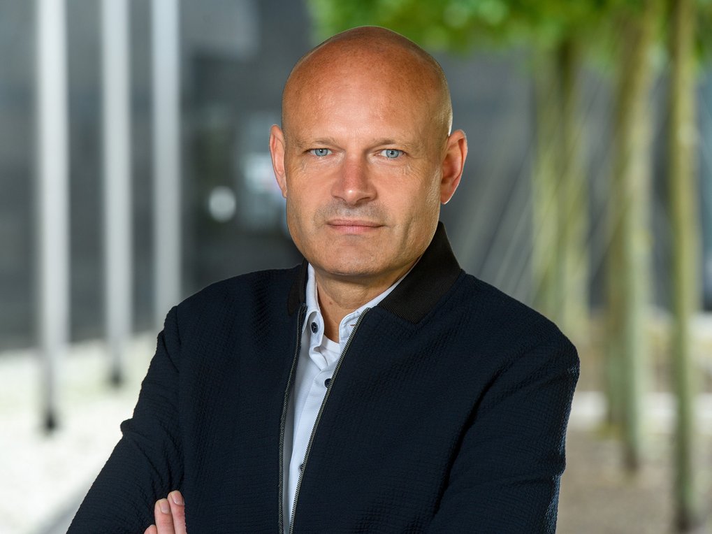 Person trägt ein weißes Hemd unter einer dunkelblauen Jacke mit strukturierter Oberfläche, steht mit verschränkten Armen vor einem unscharfen Hintergrund mit Bäumen und Glasfassade.