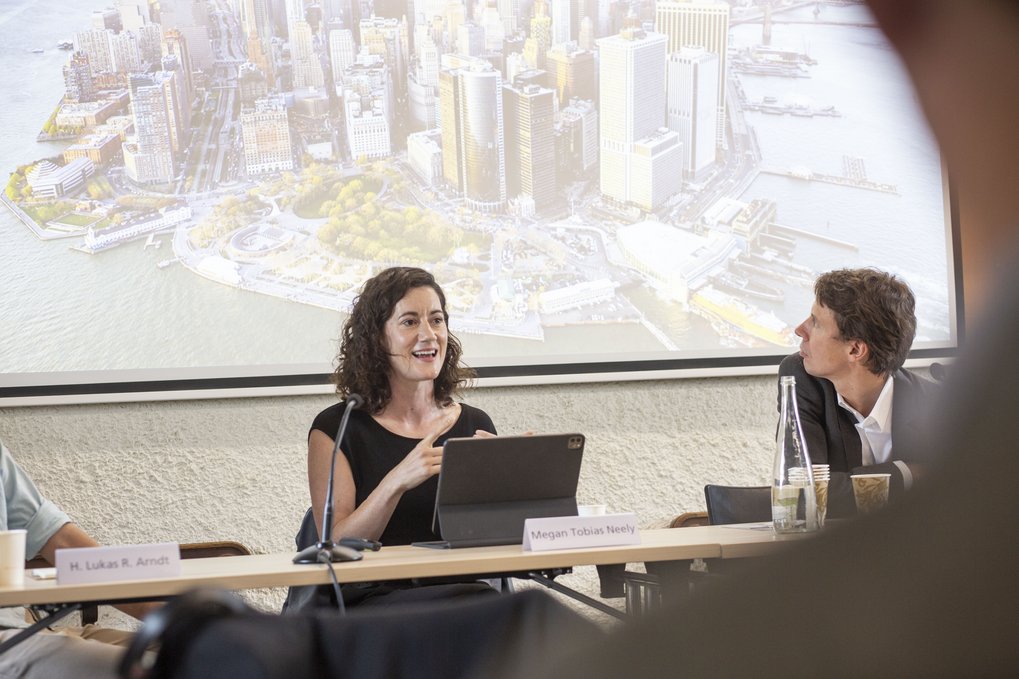 Eine Frau spricht an einem Konferenztisch, hinter ihr ist auf einer Leinwand eine Stadtlandschaft zu sehen.