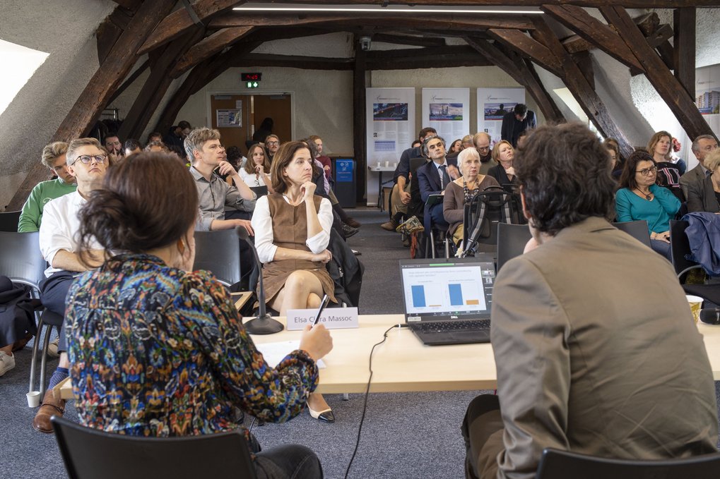 Eine Gruppe von Personen, die an einer Präsentation oder Diskussion in einem Innenraum teilnimmt, wobei der Fokus auf zwei Personen liegt, die an einem Tisch sitzen und dem Publikum zugewandt sind.