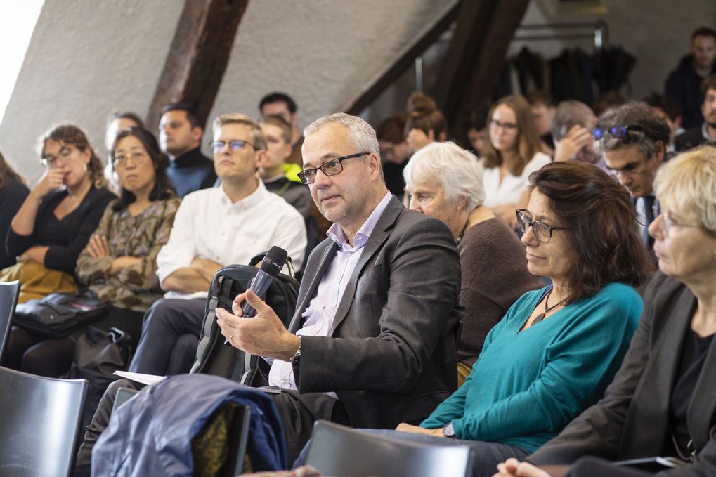 Eine Gruppe von Menschen, die während einer Diskussion oder Präsentation im Publikum sitzt. Ein Mann im Vordergrund spricht und hält ein Mikrofon in der Hand, während die anderen aufmerksam zuhören.