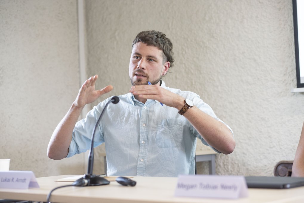 Ein Mann gestikuliert, während er an einem Tisch mit Mikrofon und Notizen spricht, während einer Diskussion oder Präsentation.