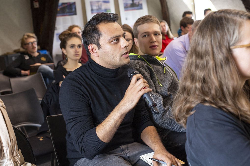 Ein Mann in einem schwarzen Pullover hält ein Mikrofon in der Hand und spricht während einer Diskussion, während andere Teilnehmer in einem Seminar oder Workshop aufmerksam zuhören.