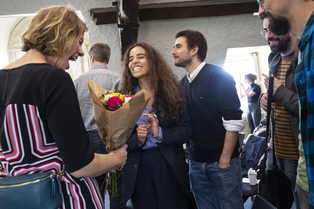 A panel discussion with several speakers at a table. One speaker, a woman with long dark hair and a colorful blouse, is actively participating and gesturing. The other panelists are seated behind her, with laptops and notes visible on the table.