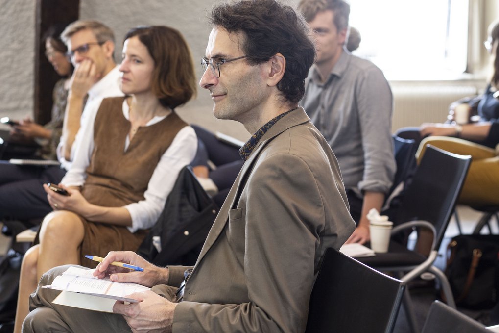 Eine Gruppe von Menschen, die in einer Besprechung oder einem Seminar sitzt, mit Fokus auf einen Mann in einem hellbraunen Anzug, der ein Buch und einen Stift in der Hand hält und sich engagiert an der Diskussion beteiligt.