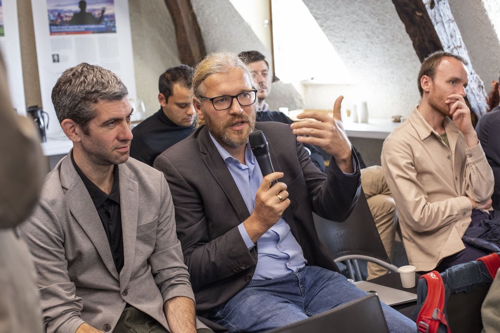 Eine Gruppe von Menschen sitzt in einem Innenraum, wobei ein Mann im Anzug in ein Mikrofon spricht und dabei gestikuliert. Die anderen hören aufmerksam zu.