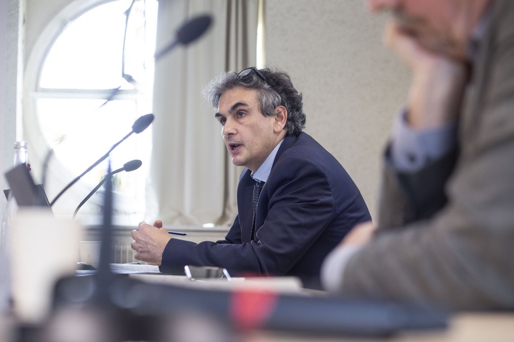 Ein Mann im Anzug spricht während einer Besprechung, im Hintergrund sind Mikrofone und ein Fenster zu sehen.