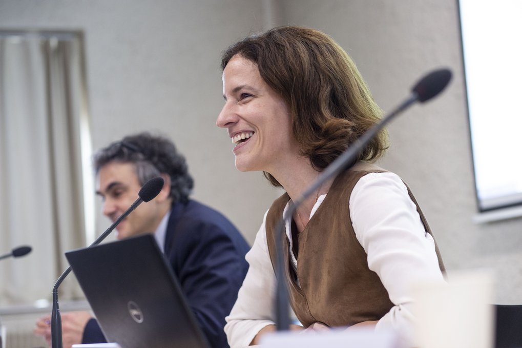Eine Frau lächelt, während sie an einer Tischdiskussion teilnimmt, mit einem Mann im Hintergrund zu ihrer Linken und einem Laptop vor ihr.