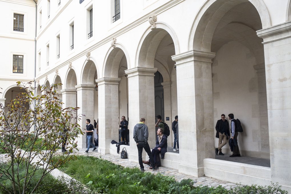 Ein Innenhof mit gewölbten Säulen, in dem Menschen sich unterhalten und in Gruppen inmitten von Grünpflanzen stehen.