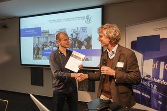 Two individuals shaking hands at a Max Planck Institute for the Study of Societies event, with a presentation slide featuring climate protest imagery.