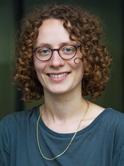 Das Porträtfoto zeigt Annika Holz, eine junge Frau mit lockigem Haar und einer Brille mit ovalen Gläsern, die Doktorandin an der IMPRS-SPCE gewesen ist.