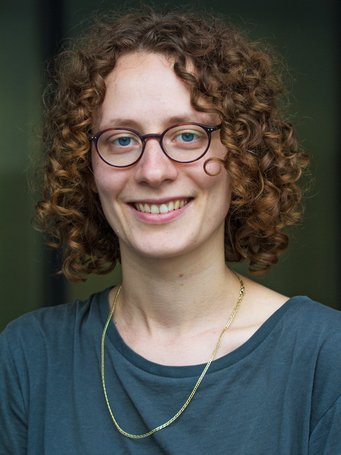 Individual with curly brown hair, wearing a simple gold necklace and a plain blue shirt, against a blurred dark background.