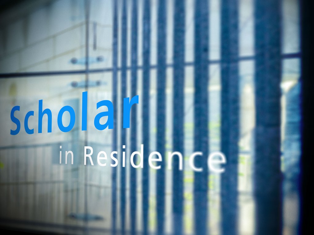 Das Bild zeigt eine reflektierende Oberfläche, auf der der Text „Scholar in Residence“ zu sehen ist.