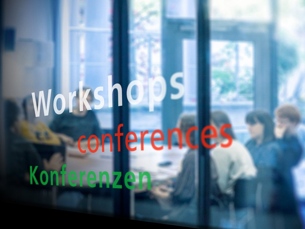 Ein unscharfes Bild, das die Wörter „Workshops“, „Konferenzen“ und „Konferenzen“ auf einer Glasoberfläche zeigt, mit Menschen, die um einen Tisch herum sitzen und im Hintergrund zu sehen sind.