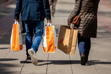 A man and a woman are walking down a city sidewalk, each carrying multiple shopping bags from different retail stores, including orange and brown bags.