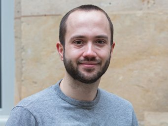 A person with short hair is wearing a gray sweater and standing in front of a beige stone wall with a window frame at the edge of the image.