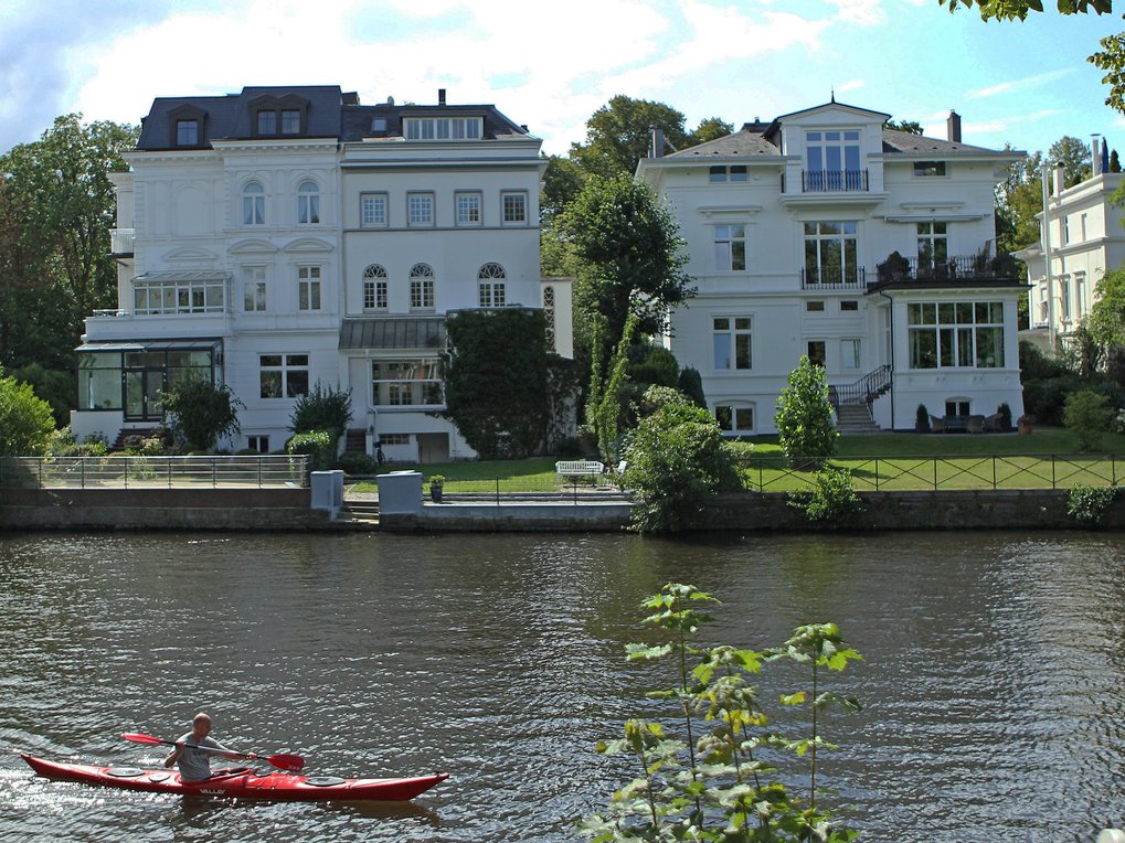 Zwei imposante, weiße Gründerzeitvillen stehen am Flussufer, während ein einzelner Kajakfahrer in einem roten Boot vorbeigleitet.