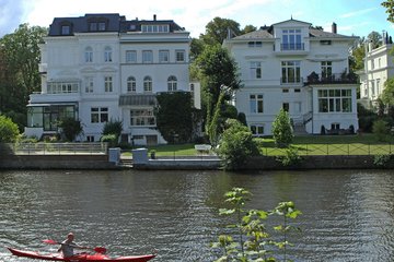 Zwei imposante, weiße Gründerzeitvillen stehen am Flussufer, während ein einzelner Kajakfahrer in einem roten Boot vorbeigleitet.