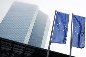 Zwei europäische Flaggen wehen vor einem hohen, modernen Hochhaus mit verglaster Fassade. Der Himmel ist neblig und grau, was eine gedämpfte Atmosphäre erzeugt.