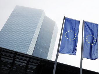 Zwar wissen Zentralbanken um ihre begrenzten Mittel bei der Inflationsbekämpfung, folgt man jedoch sich häufenden Belegen aus der Fachwelt, lassen sie die Öffentlichkeit darüber bewusst im Irrglauben. So erhalten sie ihr Image als Herrscherinnen über die Geldwertstabilität aufrecht. Zwei europäische Flaggen wehen vor einem hohen, modernen Hochhaus mit verglaster Fassade. Der Himmel ist neblig und grau, was eine gedämpfte Atmosphäre erzeugt.