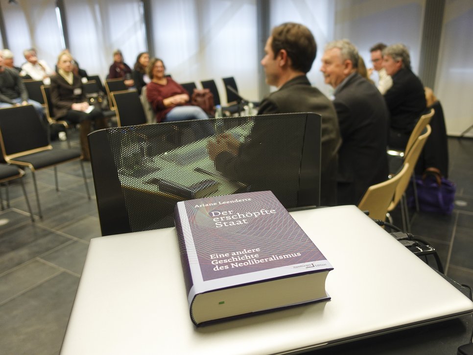 Die Fotoaufnahme zeigt eine Szene aus der Buchpräsentation zu "Der erschöpfte Staat: Eine andere Geschichte des Neoliberalismus", dem Werk von Ariane Leendertz. Im Vordergrund, auf einem Pult, ist ein Exemplar des Buches deutlich zu sehen. Die Individuen, die dem Publikum zugewandt in vorderer Reihe an Tischen sitzen, sind lediglich verschwommen zu erkennen, ebenso wie die Personen im Auditorium gegenüber.