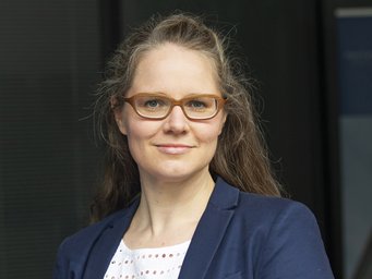 Vermutlich eine Frau mit langen, braunen Haaren, trägt einen blauen Blazer und eine weiße Bluse mit Lochmuster, vor einem dunklen Hintergrund.