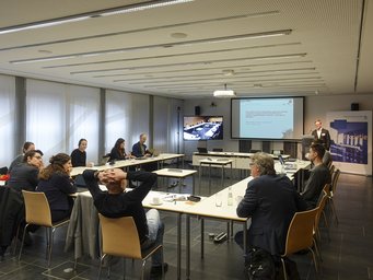 A professional meeting in a conference room, where attendees are seated around tables, observing a presentation projected on a screen with additional content visible on a monitor.