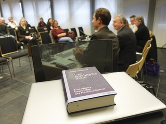 In a conference room, the book "Der erschöpfte Staat" lies on a laptop, while several attendees are seated in chairs, engaged in the event.