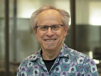An individual with gray hair wearing a shirt featuring circular green and purple patterns, situated in an office space with glass partitions.