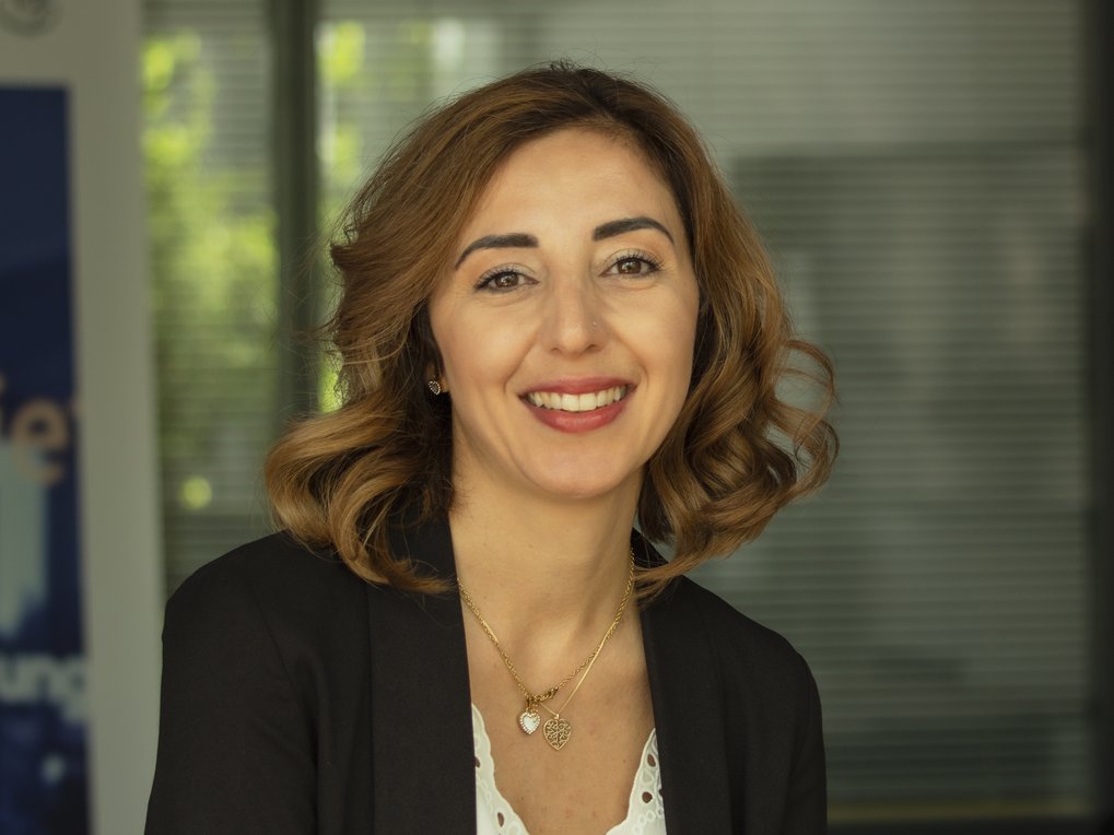 Frau mit lockigem Haar trägt einen schwarzen Blazer und zwei goldene Herzanhänger, vor einem Fenster mit Jalousien.