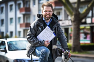 Eine Person mit Jacke und Jeans auf einem Fahrrad, hält eine Liste und steht vor einem parkenden Auto und Wohngebäuden.