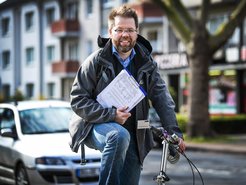 Eine Person mit Jacke und Jeans auf einem Fahrrad, hält eine Liste und steht vor einem parkenden Auto und Wohngebäuden.