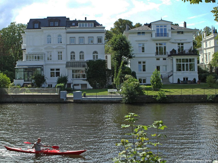Vermögende Familien als Akteure im gegenwärtigen Kapitalismus Zwei imposante, weiße Gründerzeitvillen stehen am Flussufer, während ein einzelner Kajakfahrer in einem roten Boot vorbeigleitet.
