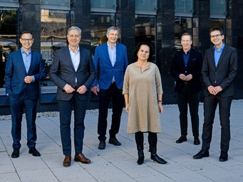 A diverse group of six individuals dressed in formal attire, positioned on a paved surface outdoors, with a modern glass-and-stone building behind them.