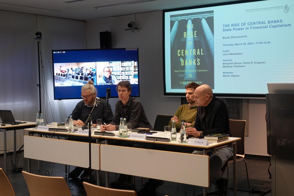 A panel discussion setup featuring several panelists seated at a table equipped with microphones, water bottles, and books. Behind them is a large screen displaying a presentation slide for a book titled "The Rise of Central Banks: State Power in Financial Capitalism" by Leon Wansleben, indicating details of the discussion event including panelists and moderator.