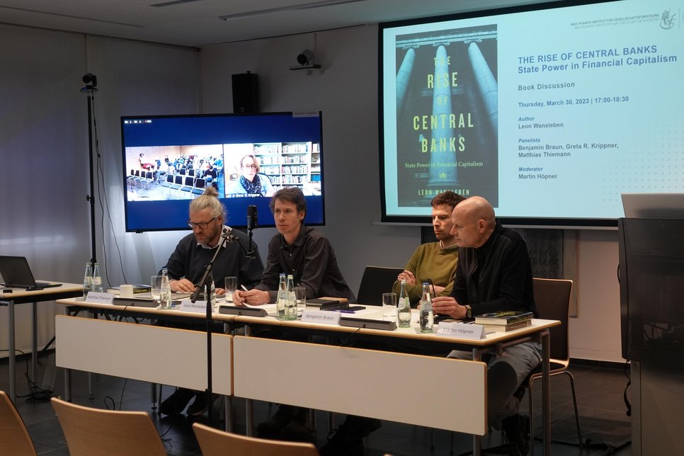 A panel discussion setup featuring several panelists seated at a table equipped with microphones, water bottles, and books. Behind them is a large screen displaying a presentation slide for a book titled "The Rise of Central Banks: State Power in Financial Capitalism" by Leon Wansleben, indicating details of the discussion event including panelists and moderator.