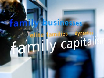 Eine Büste im Vordergrund, umgeben von reflektierenden Begriffen wie „Familienunternehmen“, „Elitefamilien“ und „dynastische Familien“ auf einer Glaswand.