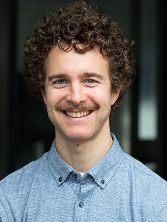 A person with curly brown hair wears a light blue button-up shirt, standing in front of a dark, out-of-focus background.