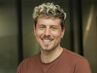 A person with curly blond hair, dressed in a brown shirt, stands in an indoor setting with a blurred background.