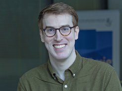 Das Porträt-Foto zeigt den Wissenschaftler Ned Crowley, der ein Stipendium des Isaac Newton Trusts (Cambridge) erhalten hat. Ganz offenbar drückt sein strahlendes Lächeln die freudige Emotion über dieses Ereignis aus. Crowley trägt ein dunkelgrünes Hemd mit Kragen, das zum Hals hin geöffnet ist. Außerdem trägt er eine Brille, womit er beabsichtigt, den Durchblick zu bewahren. Erwähnenswert ist das kurze, aber volle dunkelblonde Haar. Das Individuum vermittelt Engagement, Sympathie und Vorwärtsdrang - Eigenschaften, die ihn ganz zweifellos weit in die Welt hinaustragen werden.