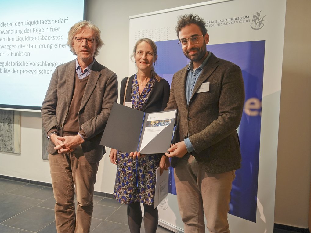 Drei Personen in formeller Kleidung präsentieren eine geöffnete Mappe mit Dokumenten, neben einem Bildschirm mit Präsentationstexten. Links ein Display mit Text über finanzielle Regeln, rechts ein Banner der Organisation.