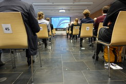 Teilnehmer sitzen in einem modernen Seminarraum auf nummerierten Stühlen, während eine Präsentation auf einer großen Leinwand zu sehen ist.