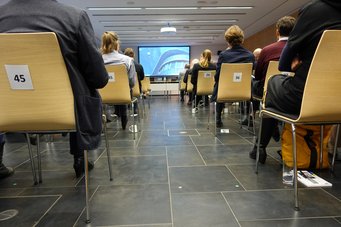 Teilnehmer sitzen in einem modernen Seminarraum auf nummerierten Stühlen, während eine Präsentation auf einer großen Leinwand zu sehen ist.