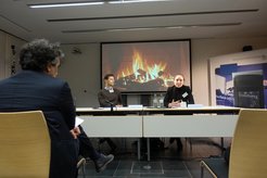Drei Personen in einem Konferenzraum, vor ihnen ein Tisch mit Mikrofonen und Wasserflaschen. Projektor zeigt Kaminfeuer. Ein Banner des Max-Planck-Instituts steht daneben.
