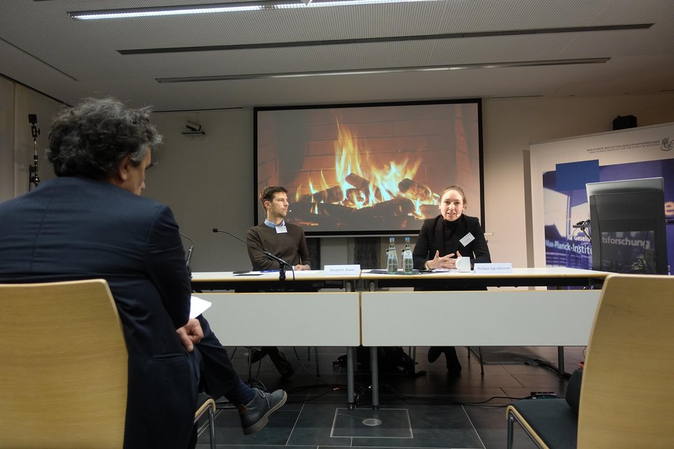 Drei Personen in einem Konferenzraum, vor ihnen ein Tisch mit Mikrofonen und Wasserflaschen. Projektor zeigt Kaminfeuer. Ein Banner des Max-Planck-Instituts steht daneben.
