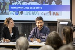 Zwei Personen an einem Konferenztisch, im Hintergrund ein großes Schild mit der Aufschrift "Klimawandel und Demokratie". Vor dem Tisch sitzt ein Publikum, das der Diskussion zuhört. Ein Rednerpult mit einem Namensschild ist ebenfalls zu sehen.