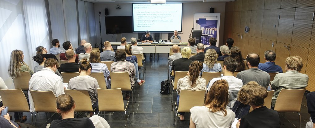Eine Gruppe von Menschen sitzt in einem modernen Konferenzraum und blickt nach vorne, wo eine Präsentation stattfindet. Zwei Redner sind am Tisch zu sehen, während die Teilnehmer aufmerksam zuhören. Im Hintergrund eine Leinwand mit projiziertem Text. Der Raum hat helle Wände und eine moderne Einrichtung.